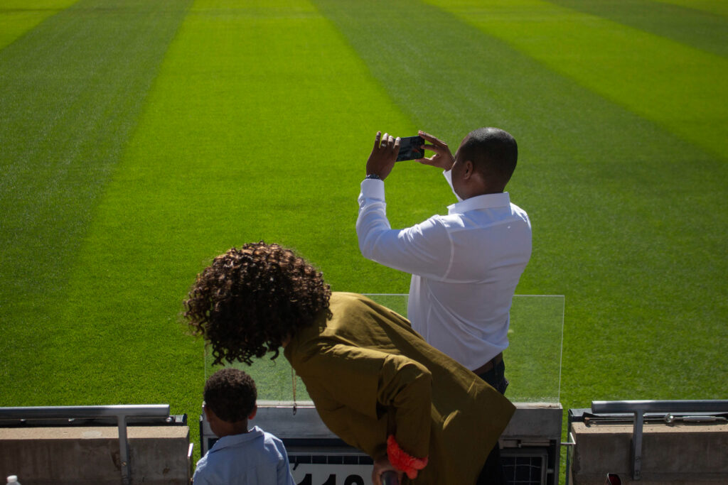 A man wearing a white shirt records on his cell phone as a woman and small child stand next to him, set against the green grass of Soldier Field.