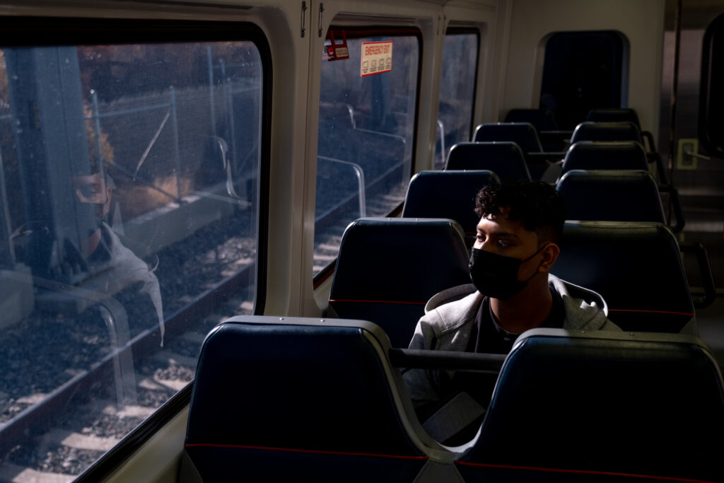 The mid-morning light shines on a young man riding the train, whose reflection looks back at him in the window.