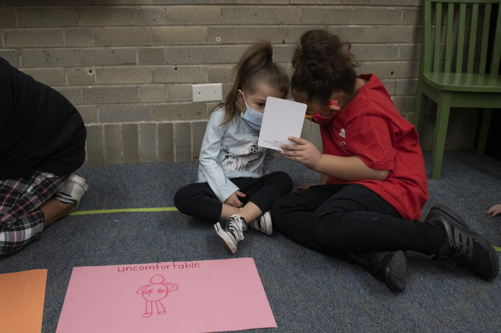 Two young girls hold up a card as they talk during a social-emotional learning lesson. They sit on a grey carpet, and there is a drawing in front of them on pink paper that says “uncomfortable” with a drawing of a person holding their abdomen.