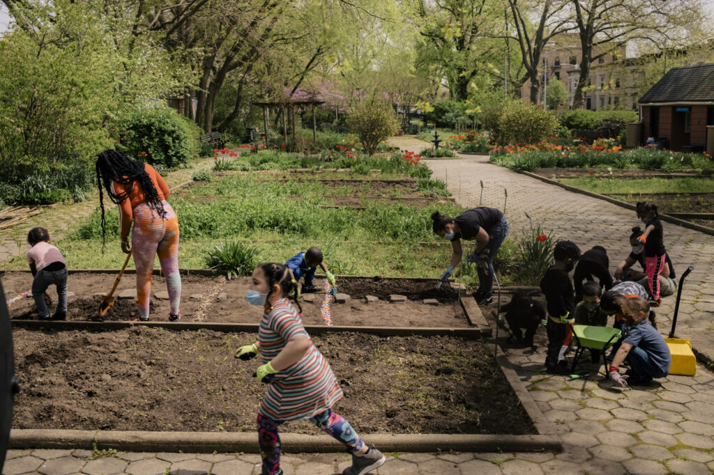 A group of kids and two women work in a community garden. A girl wearing a striped dress runs with dirt in her hands in the foreground as they work on two bare plots.