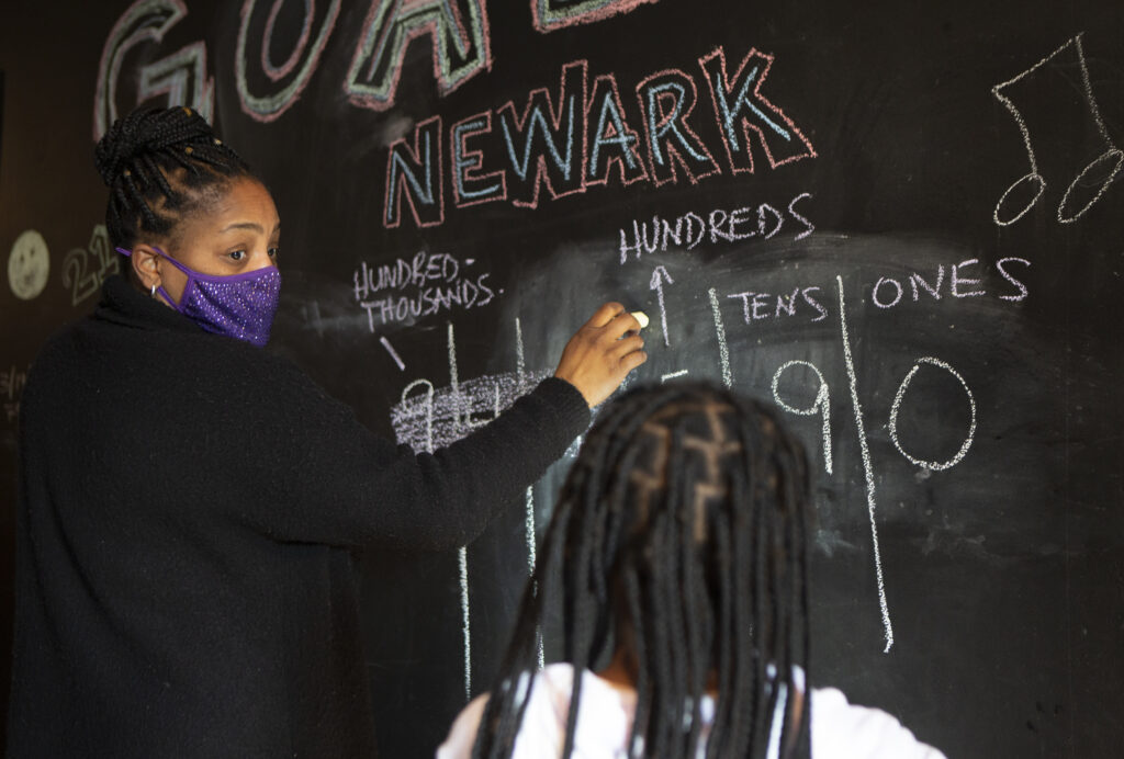 A mother, wearing a purple mask, teaches her daughter numbers on a large blackboard in their home.