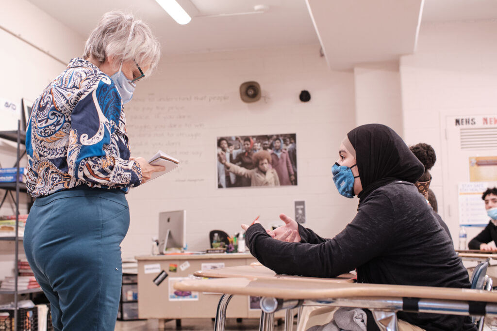 Reporter in a patterned shirt and protective mask holding a notebook while speaking with a student wearing a mask and head covering.