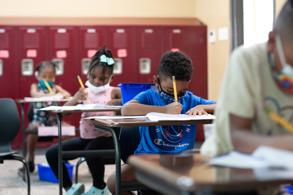 Four students sit in a row of desks in a classroom lined with red lockers. One boy wears a blue shirt and blue mask while writing on a worksheet.